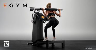 Eine Frau in Sportkleidung macht Kniebeugen an einem modernen EGYM-Trainingsgerät mit Display in einem hellen Fitnessraum.