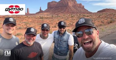 Fünf lachende Männer mit schwarzen Basecaps posieren bei sonnigem Wetter vor der roten Felsenlandschaft des Monument Valley im Südwesten der USA. Alle tragen Freizeitkleidung; im Hintergrund sind markante Tafelberge und das gym80-Logo zu sehen.