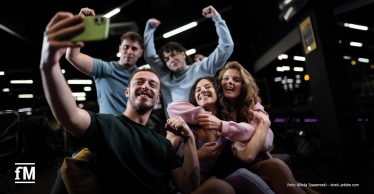 Fünf junge Erwachsene sitzen gemeinsam in einem Fitnessstudio und machen ein Gruppen-Selfie.