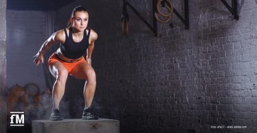 Eine sportliche Frau in Fitnesskleidung springt konzentriert auf eine Holzbox in einem Trainingsraum mit Ziegelwand.