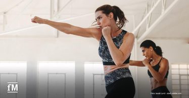 Zwei Frauen in Sportkleidung führen in einem hellen Kursraum synchron Boxbewegungen aus.