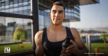 Outdoor-Trainingsfläche vor modernem Gebäude; ein sportlich gekleideter Mann steht neben einer Trainingsstation und hält einen Shaker in der Hand