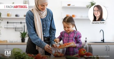 In einer hellen Küche stehen links im Vordergrund eine Frau, an ihrer Seite ein Mädchen an einer Arbeitsfläche, schneiden Tomaten auf einem Holzbrett und geben sie in eine Glasschüssel; auf der Arbeitsplatte liegen Gurke, Paprika und weiteres Gemüse, im Hintergrund sind Regale und eine Spüle zu sehen.