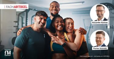 Vier Personen stehen im Vordergrund eines Fitnessstudios eng zusammen, Arme umeinander gelegt, frontal zur Kamera; im Hintergrund Trainingsgeräte und Innenraum sichtbar.