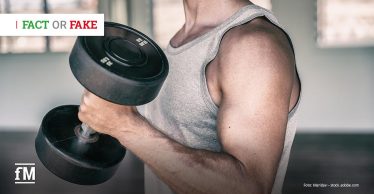 Mann in grauem Tanktop hält eine schwarze Kurzhantel im rechten Arm, Fokus auf den angespannten Oberarmmuskel in einem Fitnessraum.