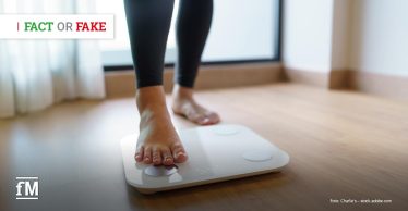 Barfüßige Person steht mit einem Fuß auf einer weißen digitalen Personenwaage auf Holzboden, der zweite Fuß befindet sich dahinter auf dem Boden
