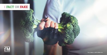 Eine Hand mit schwarzem Trainingshandschuh hält zwei Brokkoliköpfe wie eine Hantel vor hellem, unscharfem Hintergrund.