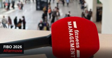 Rotes Mikrofon mit weißem „fitness MANAGEMENT“-Logo im Vordergrund, im Hintergrund unscharfe Messehalle mit gehenden Personen; links unten schwarzes Feld mit Text „FIBO 2026 Aftermovie“.