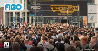 Dichte Menschenmenge vor dem Eingang der Messe Köln, im Hintergrund Glasfassade mit großen Schriftzügen „FIBO“ und „Technogym“, Banner „Welcome to FIBO“ über dem Eingang, Perspektive von hinten auf die Besucherströme