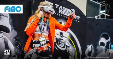 Eine blondhaarige Frau mittleren Alters trägt ein Virtual-Reality-Headset und hält zwei Controller in den Händen, während sie in Boxhaltung vor einem Messestand auf der Fitnessmesse FIBO trainiert; im Hintergrund befinden sich großflächige Grafiken mit drei weiteren Personen in VR-Ausrüstung.