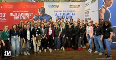 Große Gruppenaufnahme vor einer Messestand-Wand: etwa 40–50 Personen unterschiedlichen Alters und Geschlechts stehen dicht beieinander und lächeln in die Kamera