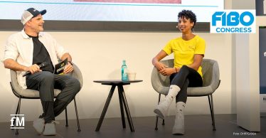 Eine Frau (Olympiasiegerin Malaika Mihambo, rechts) und ein Mann (Triathlonweltmeister Daniel Unger, links) sitzen auf Sesseln und führen ein Bühneninterview beim 'FIBO Congress', im Hintergrund ist der Anschnitt einer Powerpointpräsentation zu sehen. Die Frau trägt ein gelbes Shirt, der Moderator hält Mikrofon und Karte.