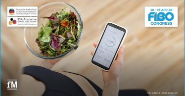 Draufsicht auf eine sitzende Person in schwarzer Sportbekleidung. In der linken Hand hält sie eine Glasschüssel mit grünem Blattsalat, in der rechten Hand ein Smartphone mit einer geöffneten Ernährungs-App, die Kalorienwerte und einen Tagesfortschritt anzeigt. Oben links sind die Logos der Deutschen Hochschule für Prävention und Gesundheitsmanagement und der BSA-Akademie eingeblendet. Oben rechts steht der Schriftzug „FIBO Congress“ mit dem Datum „16–17 Apr 26“.