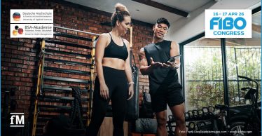 Ein Trainer und eine Sportlerin stehen in einem Fitnessstudio vor einer Backsteinwand. Der Trainer hält ein Tablet in der Hand und erklärt etwas, während im Hintergrund Trainingsgeräte wie Kettlebells und Seile zu sehen sind