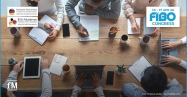 Mehrere Personen sitzen an einem großen Holztisch und arbeiten gemeinsam mit Laptops, Tablets, Notizbüchern und Kaffee.