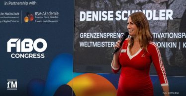 Frau in roter Adidas-Kleidung steht rechts im Vordergrund auf einer Bühne und hält ein Mikrofon in der rechten Hand. Im Hintergrund eine große Präsentationswand mit dem Schriftzug „DENISE SCHINDLER“. Links daneben eine Wand mit Logos von FIBO Congress, BSA-Akademie und DHfPG.