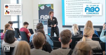 Vortragsszene in einem hellen Seminarraum: vorne eine Moderatorin mit kurzem lockigem Haar und schwarzem Anzug, die vor einem großen Bildschirm spricht; mehrere Sitzreihen mit Teilnehmern sind im Vordergrund zu sehen.