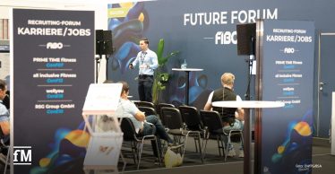 Ein Mann steht auf einer Bühne vor einem 'Future Forum'-Hintergrund auf der FIBO und spricht zu einem Publikum. Im Vordergrund stehen zwei Aufsteller mit Informationen zum Recruiting-Forum 'Karriere/Jobs' mit Unternehmenslogos.