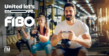 Drei Personen trainieren mit Kettlebells in einem Fitnessstudio. Im Vordergrund hält ein lächelnder Mann eine schwarze Kettlebell. Links stehen groß die Schriftzüge „United let’s move.“ und „FIBO“.