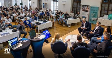 Großer Veranstaltungssaal mit mehreren Reihen besetzter Tische. Im Vordergrund sitzen Teilnehmende einer Podiumsdiskussion in Sesseln um kleine Tische. Auf Monitoren steht „FIT FORWARD Branchenforum 2025“.