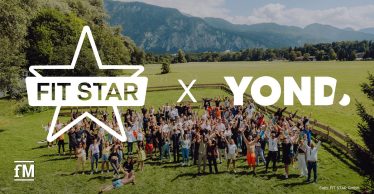 Eine große Gruppe von Menschen steht auf einer grünen Wiese mit erhobenen Armen, im Hintergrund Berge und Bäume. Im Vordergrund die Logos von FIT STAR und YOND.