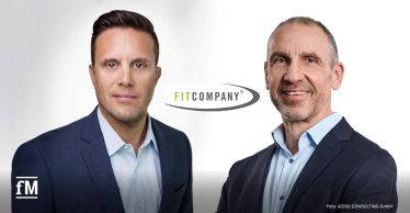 Zwei Männer in blauen Anzügen stehen vor hellem Hintergrund mit dem FITCOMPANY-Logo in der Mitte.