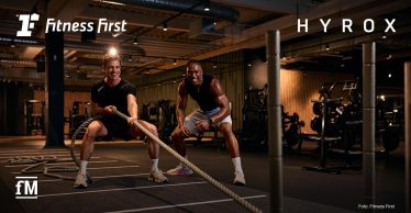 Zwei Männer trainieren mit Battle Ropes in einem Fitnessstudio; im Hintergrund Fitnessgeräte, Logos von Fitness First und HYROX eingeblendet.