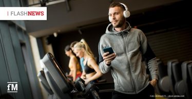 Mann mit grauem Hoodie und Kopfhörern läuft auf einem Laufband im Fitnessstudio und hält ein Smartphone in der Hand.