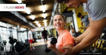 Eine lächelnde Frau im Fitnessstudio hält eine Kurzhantel, während ein Trainer ihr beim Schultertraining hilft; im Hintergrund sind weitere Sportgeräte und trainierende Personen unscharf zu sehen.