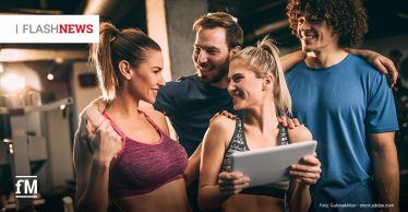 Vier junge Erwachsene in Sportkleidung stehen eng beieinander in einem Fitnessstudio, lächeln sich an und schauen auf ein Tablet; oben links steht „FLASHNEWS“.