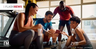 Vier lachende Fitnesssportler sitzen nach dem Training auf dem Boden eines Fitnessstudios und schauen gemeinsam auf ein Smartphone; neben ihnen stehen Trinkflaschen, im Hintergrund sind Laufbänder und große Fenster zu sehen.