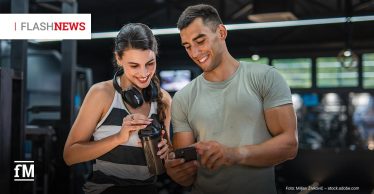 Zwei junge Erwachsene im Fitnessstudio lächeln und schauen gemeinsam auf ein Smartphone; die Frau trägt Kopfhörer um den Hals und hält einen Shaker. Oben links steht „FLASH NEWS“.