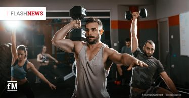 Muskelöser Mann im Fitnessstudio hebt eine Kurzhantel über den Kopf; im Hintergrund trainieren weitere Personen mit Gewichten.