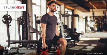 Im Vordergrund rechts trainiert ein Mann in schwarzer Sportkleidung in einem Fitnessstudio mit zwei Kettlebells in den Händen. Sein linkes Bein steht angewinkelt auf einer flachen Trainingsbank, sein rechtes Bein steht am Boden. Im Hintergrund links stehen Langhantelablagen vor hohen Fenstern, rechts liegen weitere Trainingsgeräte unscharf im Raum.