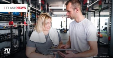 Eine Frau in Sportkleidung spricht mit einem Trainer, der ein Tablet hält. Beide blicken auf den Bildschirm. Im Hintergrund sind Krafttrainingsgeräte und Trainingszubehör zu sehen. Links oben ist das Logo „FLASHNEWS“ eingeblendet.