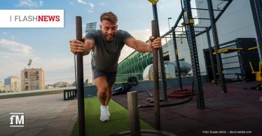 Mann schiebt im Außenbereich eines Fitnessstudios einen Gewichtsschlitten über Kunstrasen und blickt lächelnd nach vorn. Oben links steht „FLASH NEWS“, im Hintergrund sind Trainingsgeräte und Gebäude zu sehen.
