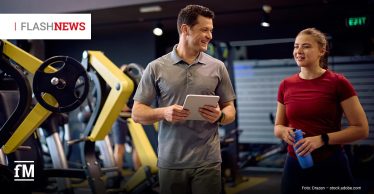 Ein Trainer mit Tablet spricht lächelnd mit einer Sportlerin, die eine Trinkflasche hält. Die beiden stehen zwischen Kraftgeräten in einem Fitnessstudio.