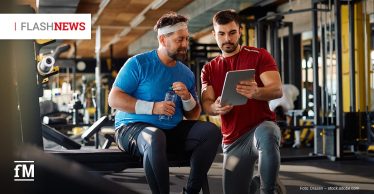 Zwei Männer in Sportkleidung sitzen in einem Fitnessstudio. Einer hält eine Wasserflasche, während der andere ihm etwas auf einem Tablet zeigt. Links oben steht „FLASHNEWS“.