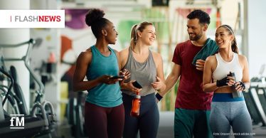 Vier lachende Personen in Sportkleidung stehen nach dem Training gemeinsam in einem Fitnessstudio. Zwei halten Trinkflaschen. Oben links steht „FLASHNEWS“.