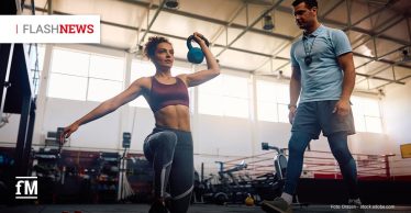 Eine Frau führt mit einer Kettlebell eine Ausfallschrittübung aus, während ein Trainer sie in einem Fitnessstudio beobachtet und anleitet.