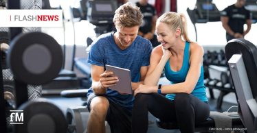Ein sportlich gekleideter Mann und eine Frau sitzen im Fitnessstudio nebeneinander auf einer Hantelbank und schauen gemeinsam lächelnd auf ein Tablet. Im Hintergrund sind Fitnessgeräte und weitere trainierende Personen zu sehen.