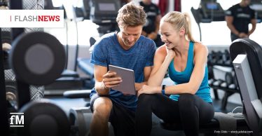 Ein lächelnder Mann und eine Frau in Sportkleidung sitzen nebeneinander im Fitnessstudio und schauen gemeinsam auf ein Tablet. Im Hintergrund sind Trainingsgeräte zu sehen. Oben links steht „FLASHNEWS“.