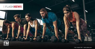 Fünf junge sportliche Personen, drei Frauen und zwei Männer, trainieren im Fitnessstudio in Liegestützposition auf Kurzhanteln. Oben links steht „FLASHNEWS“, unten links das Logo