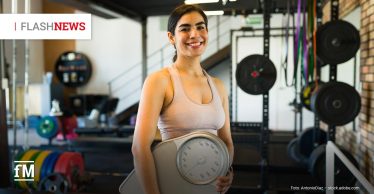 Eine junge Frau steht lächelnd in einem Fitnessstudio und hält eine große analoge Waage vor ihrem Oberkörper. Im Hintergrund sind Hanteln, Gewichtsscheiben und Sportgeräte sichtbar.