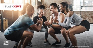 Fünf junge Erwachsene trainieren lachend in einem Fitnessstudio. Sie stehen sich paarweise gegenüber und machen gemeinsam Kniebeugen.