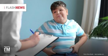 Ein übergewichtiger Junge sitzt mit beiden Händen am Bauch vor einer Ärztin oder einem Arzt (es ist nur der rechte Arm im weißen Kittel zu sehen) mit Klemmbrett, die Notizen macht.