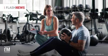 Eine Frau in türkisfarbener Sportkleidung kniet mit Klemmbrett vor einem Mann, der im Fitnessstudio mit einem Medizinball trainiert. Beide lächeln einander an.