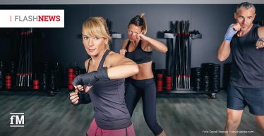 Drei Personen in Sportkleidung trainieren in einem Fitnessstudio gemeinsam eine Kampfsportübung mit Schlagbewegungen vor gestapelten Hanteln und Stange.