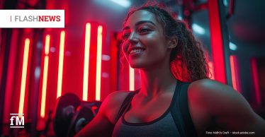 Eine junge Frau mit Sportkleidung trainiert an Hanteln in einem Fitnessstudio mit roter Beleuchtung. Sie lächelt und blickt nach vorne. Links unten befindet sich das fM-Logo und oben in der Ecke der Reiter 'Flashnews', der erste Teil grau, der zweite rot.