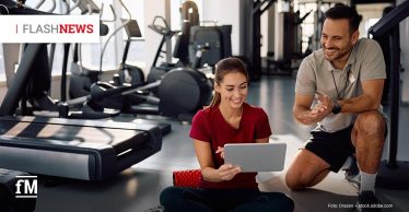 Frau in rotem Sportshirt sitzt im Fitnessstudio auf einer Matte und schaut lächelnd auf ein Tablet; daneben kniet ein männlicher Trainer in grauem Polo und schwarzer Shorts.